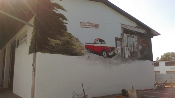 Eine Aussenwand einer Autofirma wurde in eine Tankstelle verwandelt. Ein rotes altes Auto steht an einer weißen Zapfsäule . Ein Bretterverschlag ist die Tankstelle in brauntönen. Eingerahmt links und rechts mit grünen Baumen