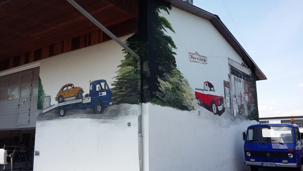 die Aussenwand ist mit einem Abschlepper in blau auf den ein Vw-Käfer Auto steht. Die Bäume sind in brün gehalten der Hintergrund ist hell