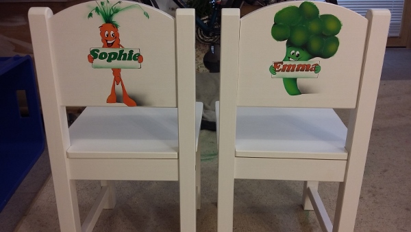 zwei weiße Kinderstühle sind mit airbrush und malen kreativ gestaltet. Die Rückseiten der Stühle sind mit einmal einer orangen Karotte die lacht und ein Schild hält mit dem Namen Sopghie in grün gestaltet. Der andere Stuhl ist mit einem grünen Brokolie der ein Schild mit dem Namen Emma hält und der in orange ist.