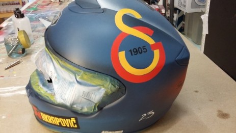 Motorradhelm in blauer Farbe als Grundfläche darauf ist in gelb und rot ein Fußballlogo angebracht auf der linken Seite.  Am Kinn ist auf dem Helm auch ein Logo in gelb und rot angebracht umfasst mit schwarz