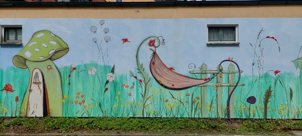 Airbrush und Malerei an einer Kindergartenaussenwand. in grün gehalten mit einem Haus als Pilz  sowie eine Hängematte gealten von zwei Blumenstielen in grün mit roten Blumen