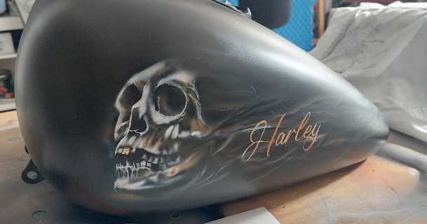 ein schwarzer Motorradtank wurde mit einem Totenkopf und einem Schriftzug geairbrusht. in weiß,schwarz und brauntönen. ein Zahn wurde mit Blattgold versehen