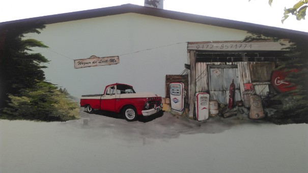 Eine Aussenwand einer Autofirma wurde in eine Tankstelle verwandelt. Ein rotes altes Auto steht an einer weißen Zapfsäule . Ein Bretterverschlag ist die Tankstelle in brauntönen. Eingerahmt links und rechts mit grünen Baumen.